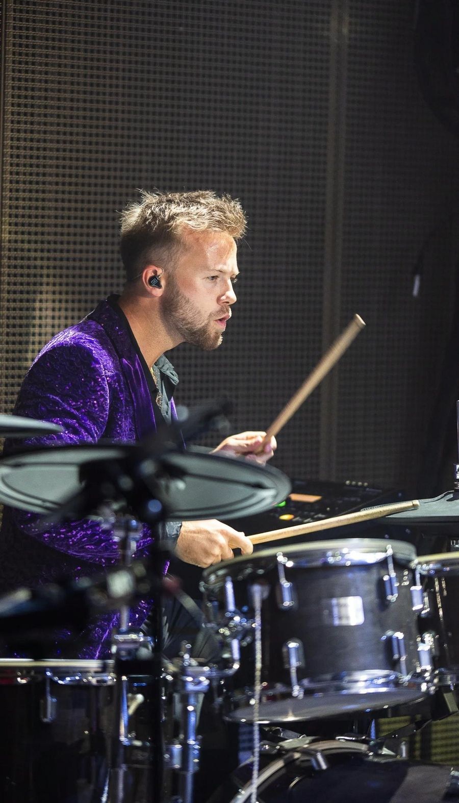 Oli playing drums on stage in a purple sequinned jacket, wearing in-ear monitors