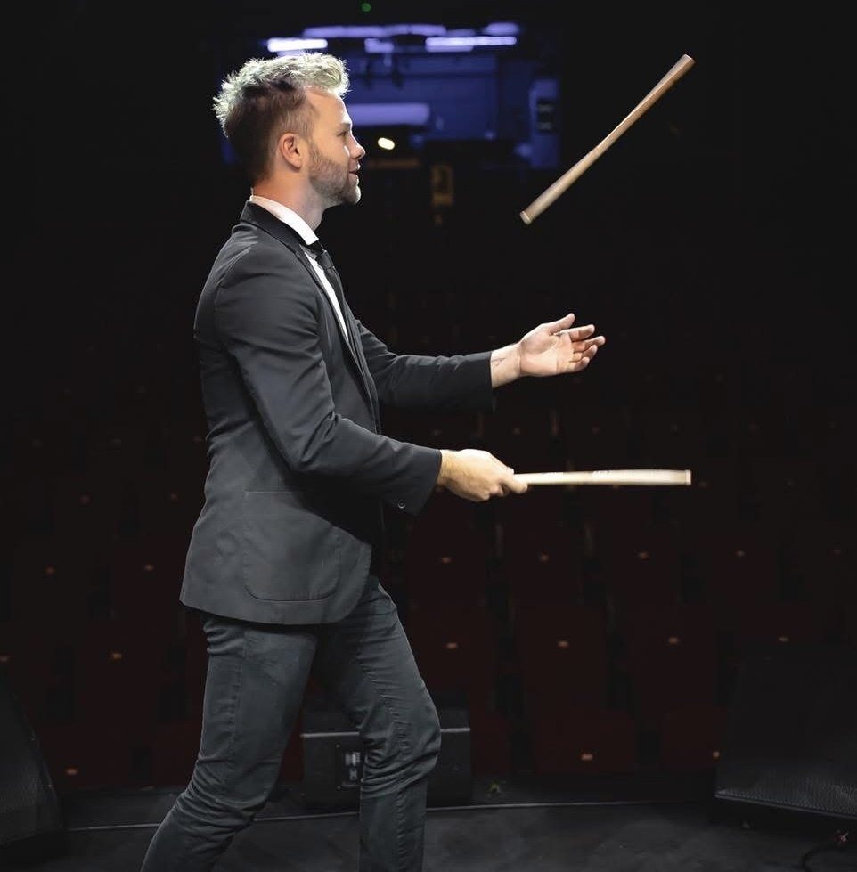 Oli Edwards in a suit on a dark stage, tossing a drumstick