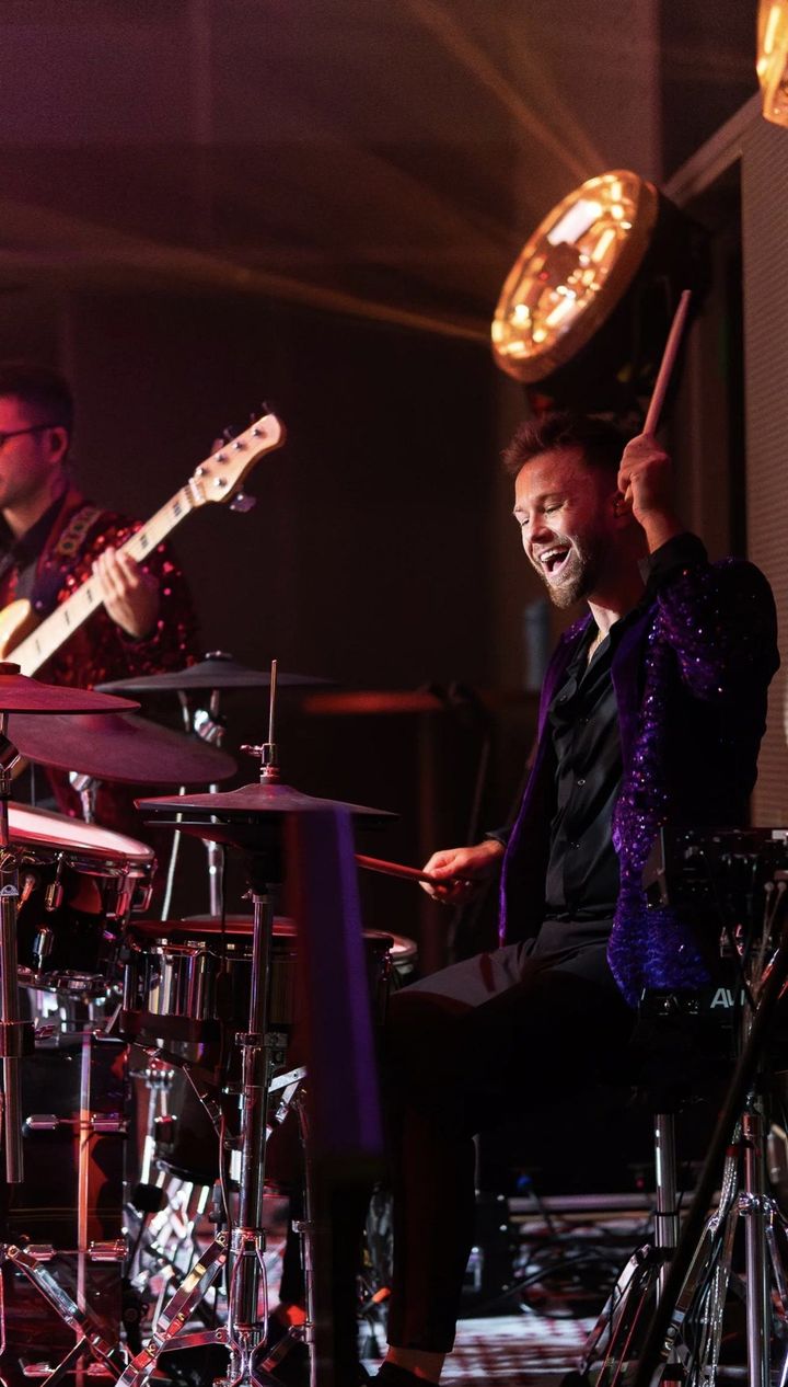 Oli laughing mid-song at the kit in a purple sequinned jacket, bassist beside him