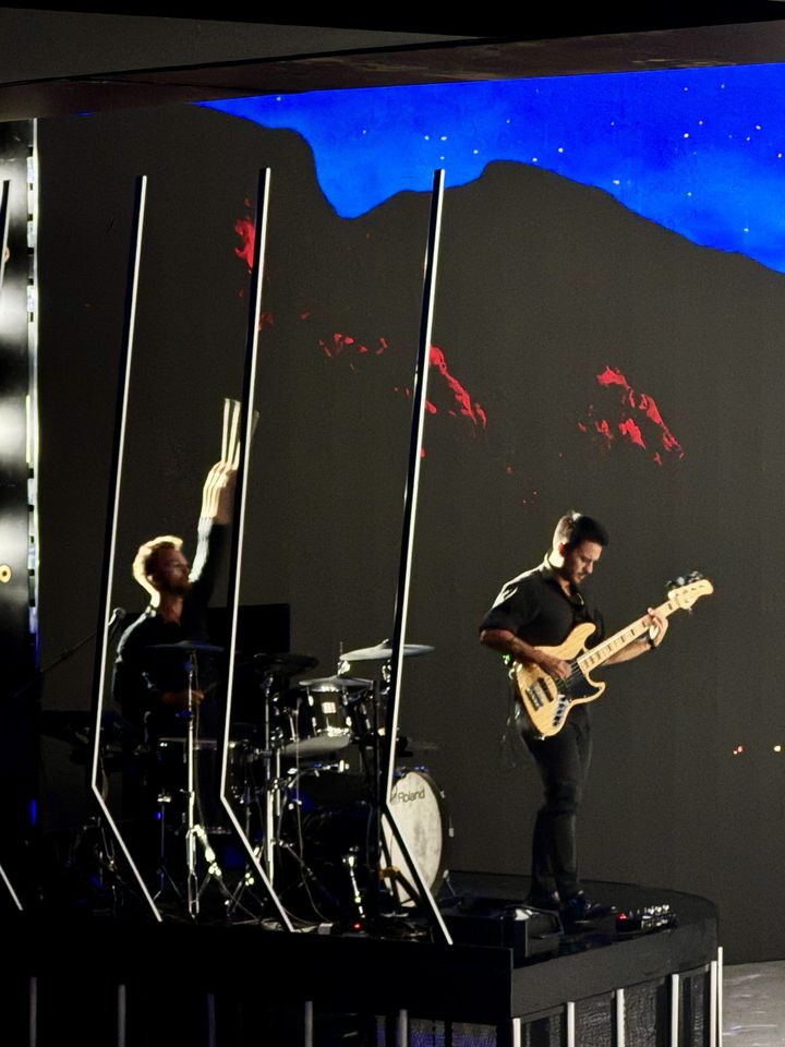 Oli at the kit behind drum screens with the bassist beside him, under a blue-lit video wall